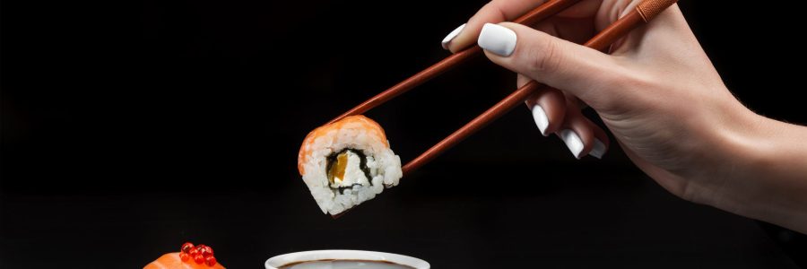 Hand holds sushi over bowl with soy sauce on black table.