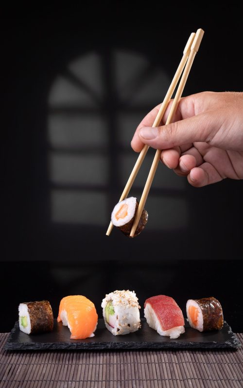 Hand picking sushi variety with chopsticks on dark background. Vertical format.